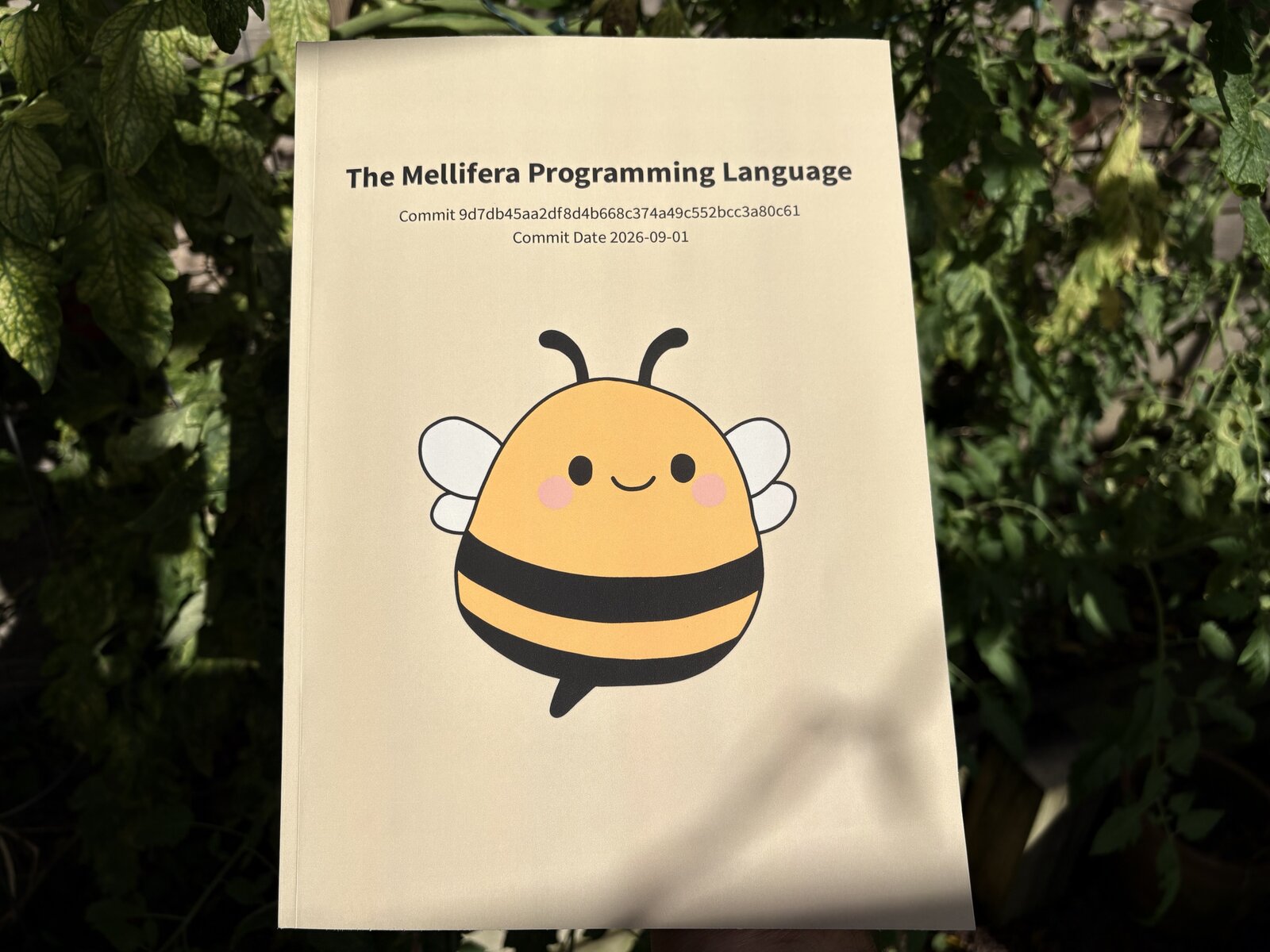 Photograph of a paperback book titled "The Mellifera Programming Language" held outside in the sunlight in front of some tomato plants. The book is 8.5x11 inches with about an inch (~300 pages) of depth, has a plain tan background, and features a cover image of Melli, a rotund cute looking cartoon bee with tiny wings who is smiling. The cover features subtitles with the commit SHA (9d7db45aa2df8d4b668c374a49c552bcc3a80c61) and the commit date (2026-09-01) used for the source code of the book.