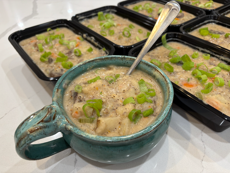 A green ceramic soup bowl filled with a mixture of potatoes, mushrooms, celery, carrots, and other vegetables. The top mixture is covered in a light dusting of freshly cracked black pepper and freshly sliced green onions. Behind the bowl sits seven to-go containers filled with the same mixture.