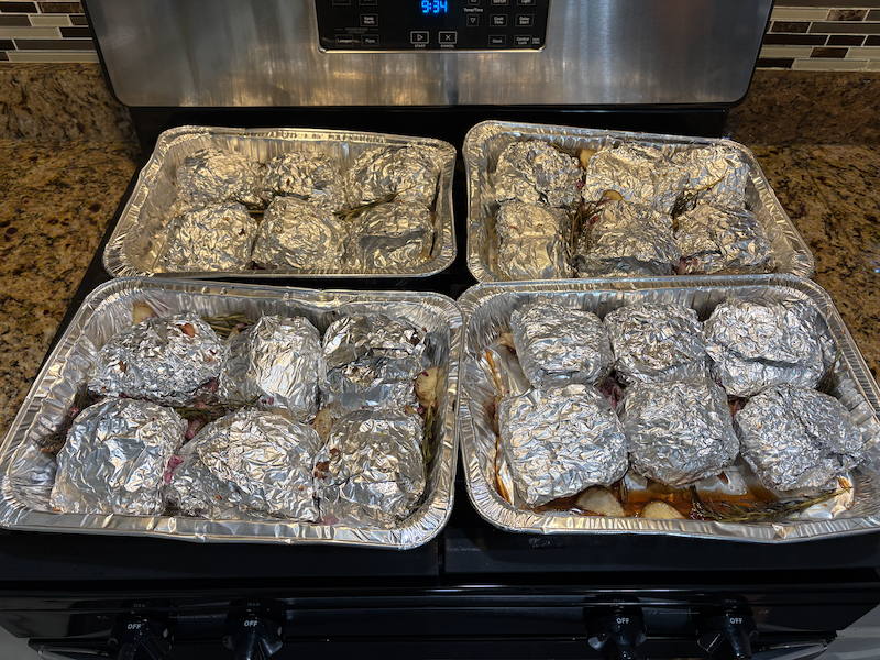 Four disposable baking trays sitting on top of an oven, each containing six foil-wrapped mixtures of meat and/or veggies. The trays are also filled with the delicious-looking juices let go by the mixtures during baking, as well as extra potatoes and rosemary that were baked alongside the wrapped mixtures.