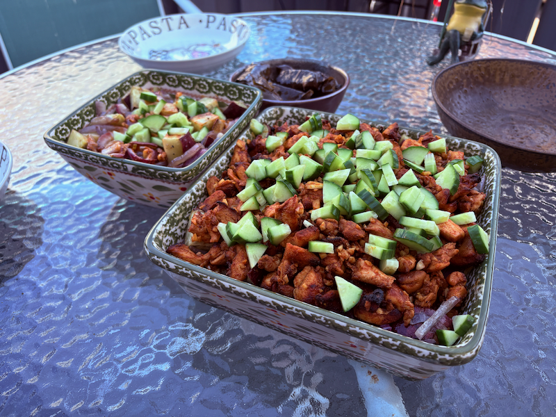 Two square-shaped ceramic bowls, one small on the left and one large on the right, sitting outside on glass table. The small bowl on the left contains a visible mixture of roasted red onion, Japanese sweet potato, and cucumber. The large bowl on the left contains a visible mixture of red onion, cucumber, and ~1 inch pieces of baked chicken covered in a spice mixture. The ends of the chicken appear to be slightly charred, as if the chicken was in the oven just long enough to fully bake without burning. In the small bowl, the vegetables are mixed together with the cucumber sitting atop the dish. In the large bowl, there chicken forms a layer that almost completely obscures the vegetables, except for the cucumber which is once again sitting atop the dish. Next to both dishes there is a small round ceramic bowl containing several dolma.